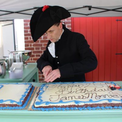 Man dressed as James Monroe, hat and all cutting a big sheet cake saying "Happy 259th Birthday James Monroe!"
